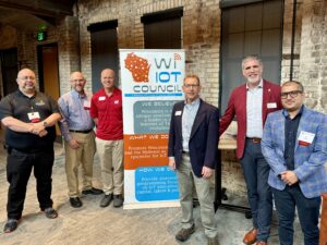 Six men in suits are standing by the Wisconsin IoT banner, smiling.